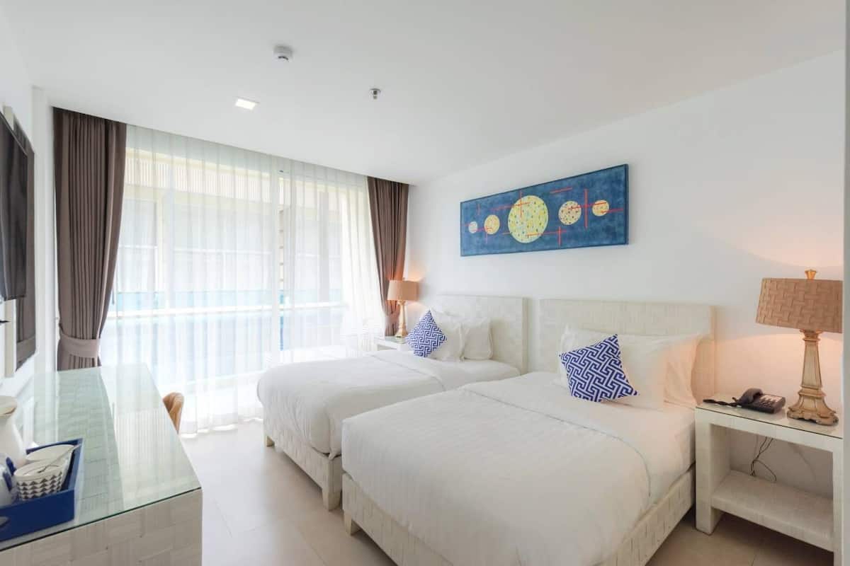 Una habitación de hotel con dos camas, un escritorio de cristal, una lámpara y un cuadro en la pared.
