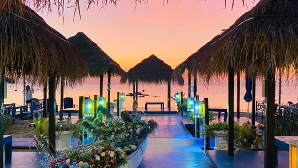 Un restaurante en la playa con sombrillas de paja, mesas al aire libre y vista al mar al atardecer.