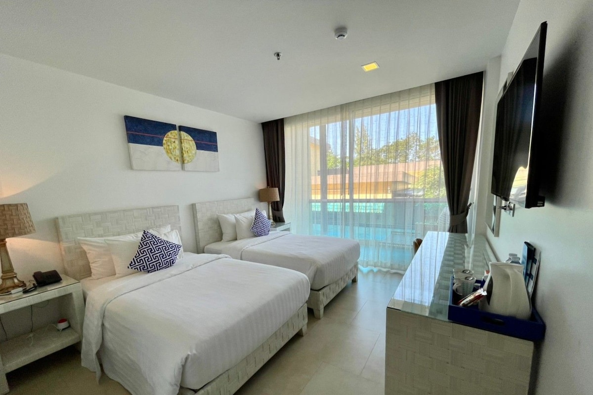 A hotel room with two beds, a desk, a lamp, a painting, and a view of the outdoors.