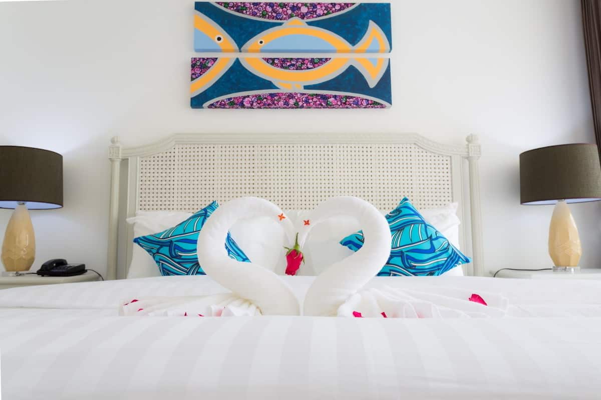 Habitación de hotel con cama, ropa de cama blanca, cabecera decorada con cabeza de cisne, un cuadro de peces y dos lámparas.