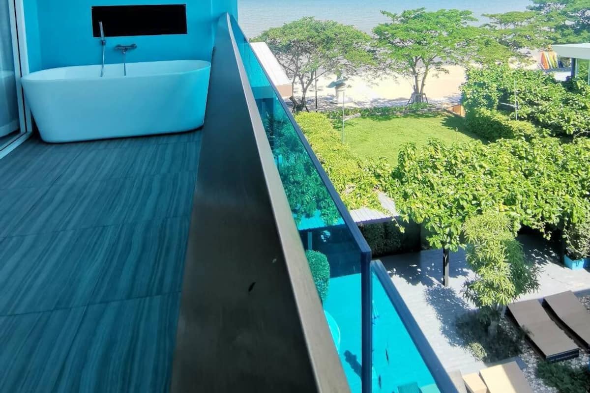 Un balcón moderno con una bañera blanca, barandilla de vidrio y vista al mar y vegetación.
