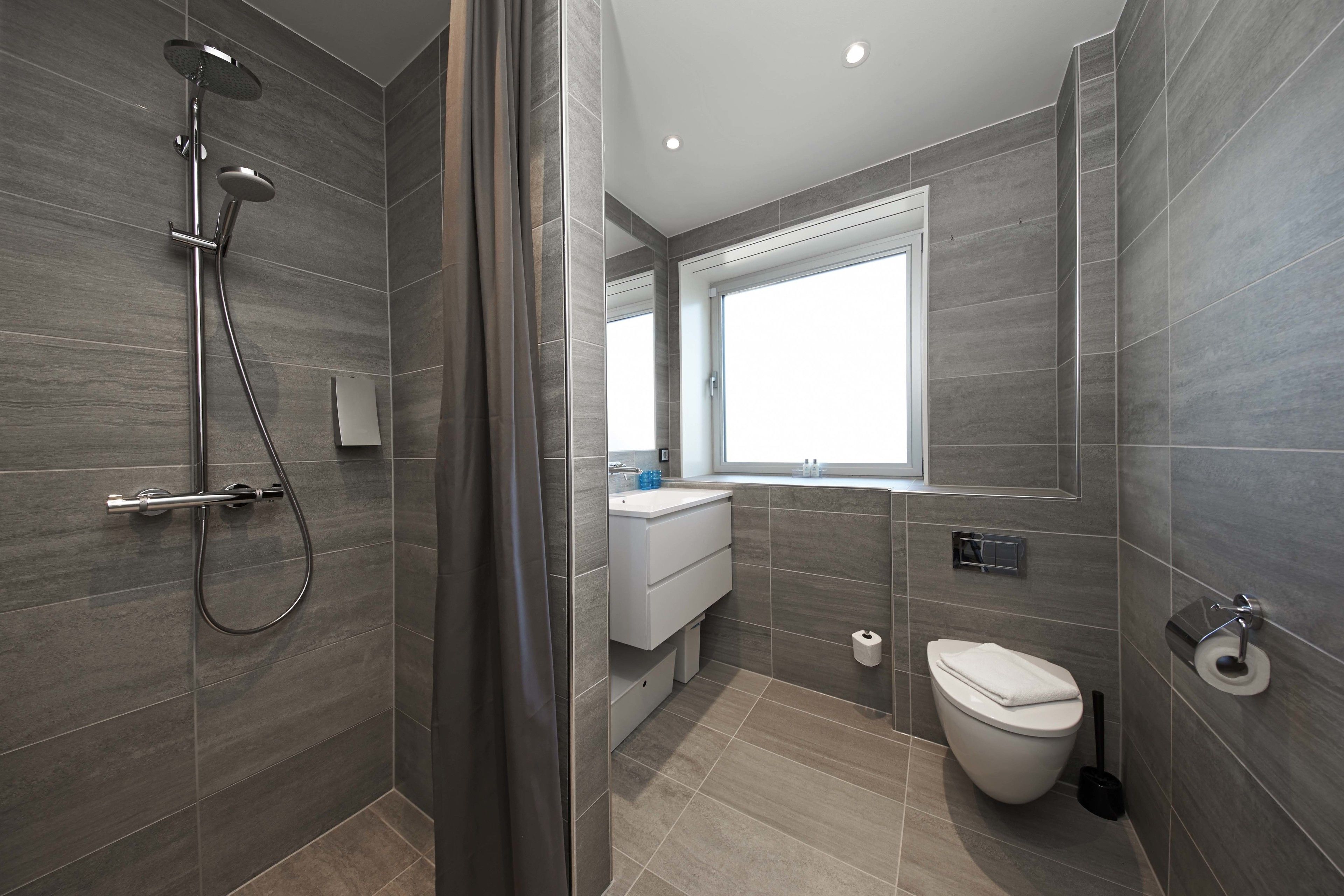 A modern bathroom with a shower, toilet, and sink, featuring grey tiles and a window.