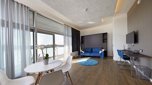 Quarto de hotel moderno com área de refeições, sofá, televisão e uma grande janela com cortinas.