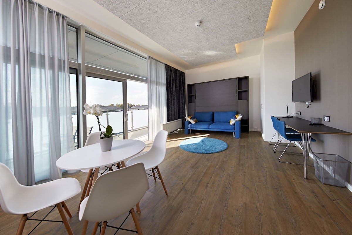 Quarto de hotel moderno com área de refeições, sofá, televisão e uma grande janela com cortinas.