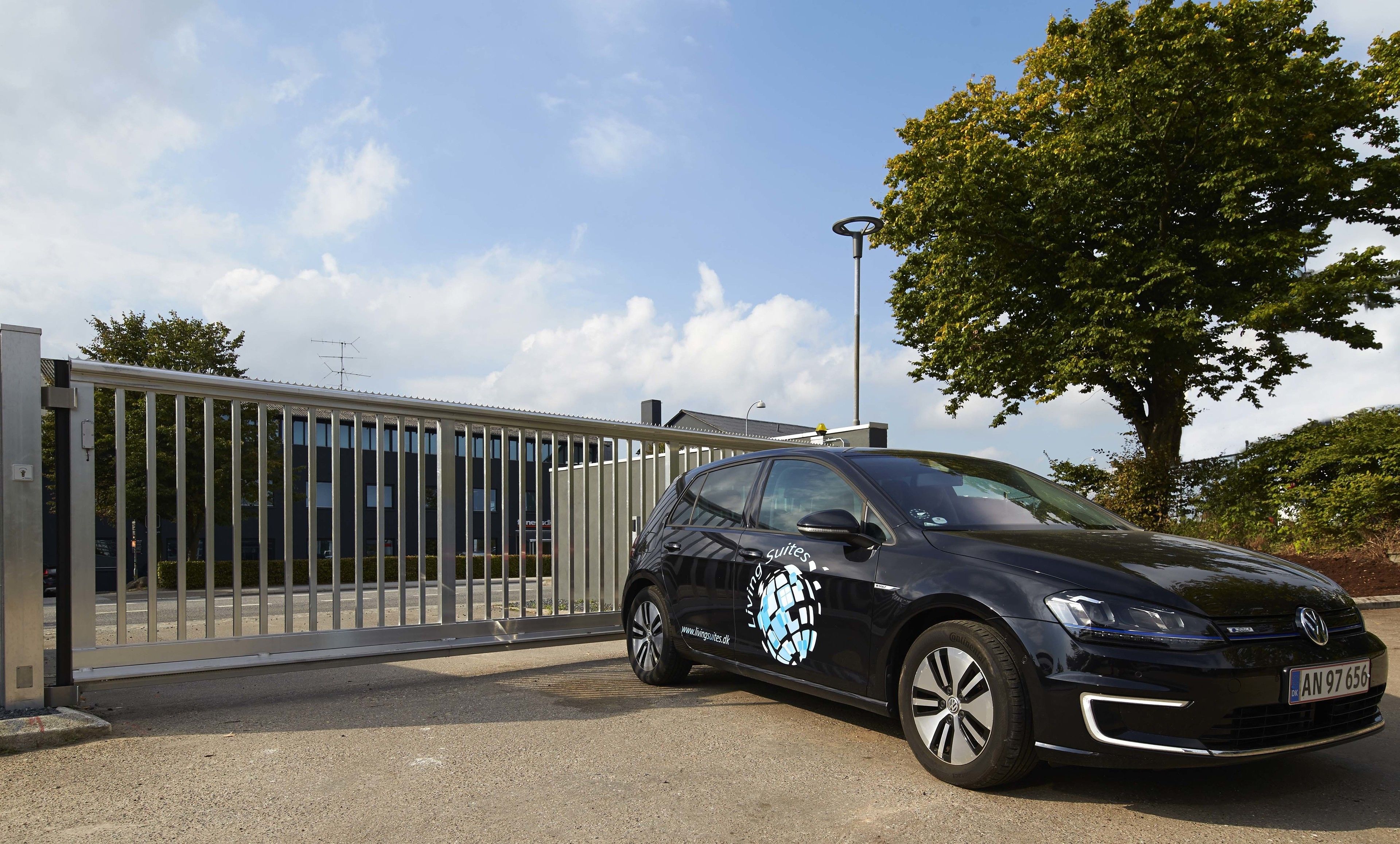 Une voiture noire est garée devant un immeuble entouré d’une grille en métal, avec un arbre en arrière-plan.