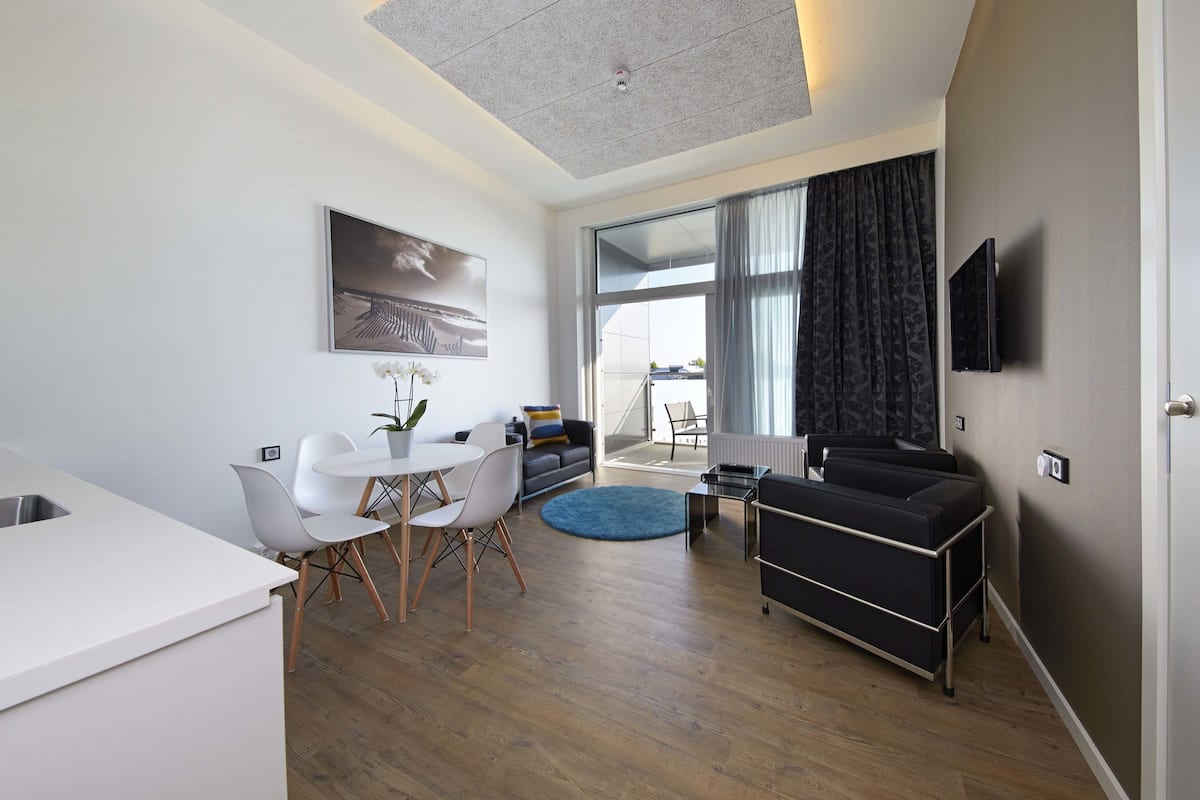 Uma sala de estar moderna com um sofá, uma área de jantar com uma mesa e cadeiras brancas, uma cozinha com bancada branca e uma janela grande com cortinas.