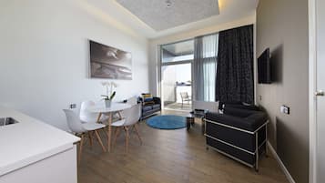 A modern living room with a sofa, a dining area with a white table and chairs, a kitchen area with a white countertop, and a large window with curtains.