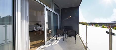 A modern balcony with a glass door leading to a kitchen area, a dining table with chairs, and a view of the cityscape.