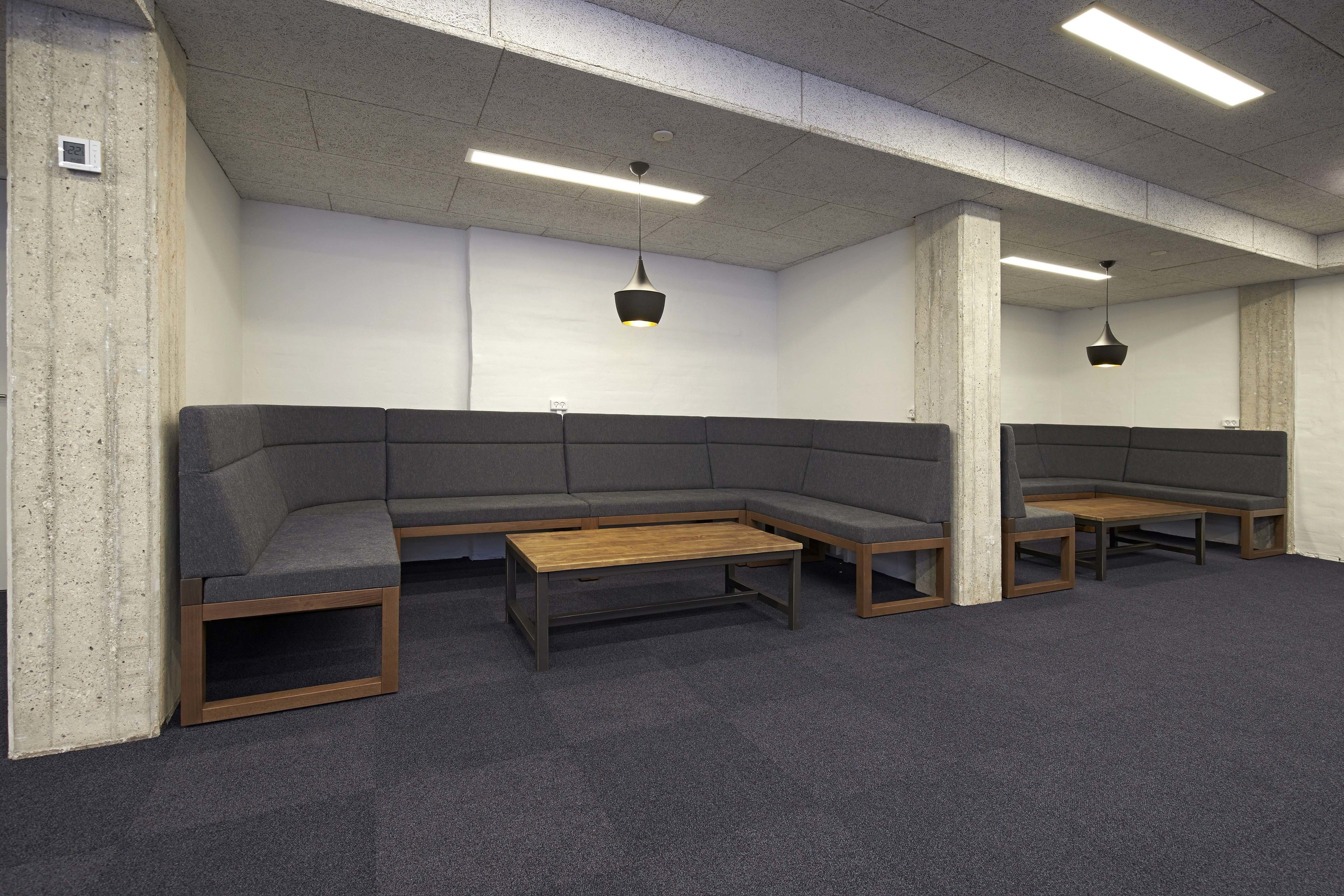 Un espace d’attente avec un canapé d’angle gris, des tables basses en bois et des luminaires suspendus.