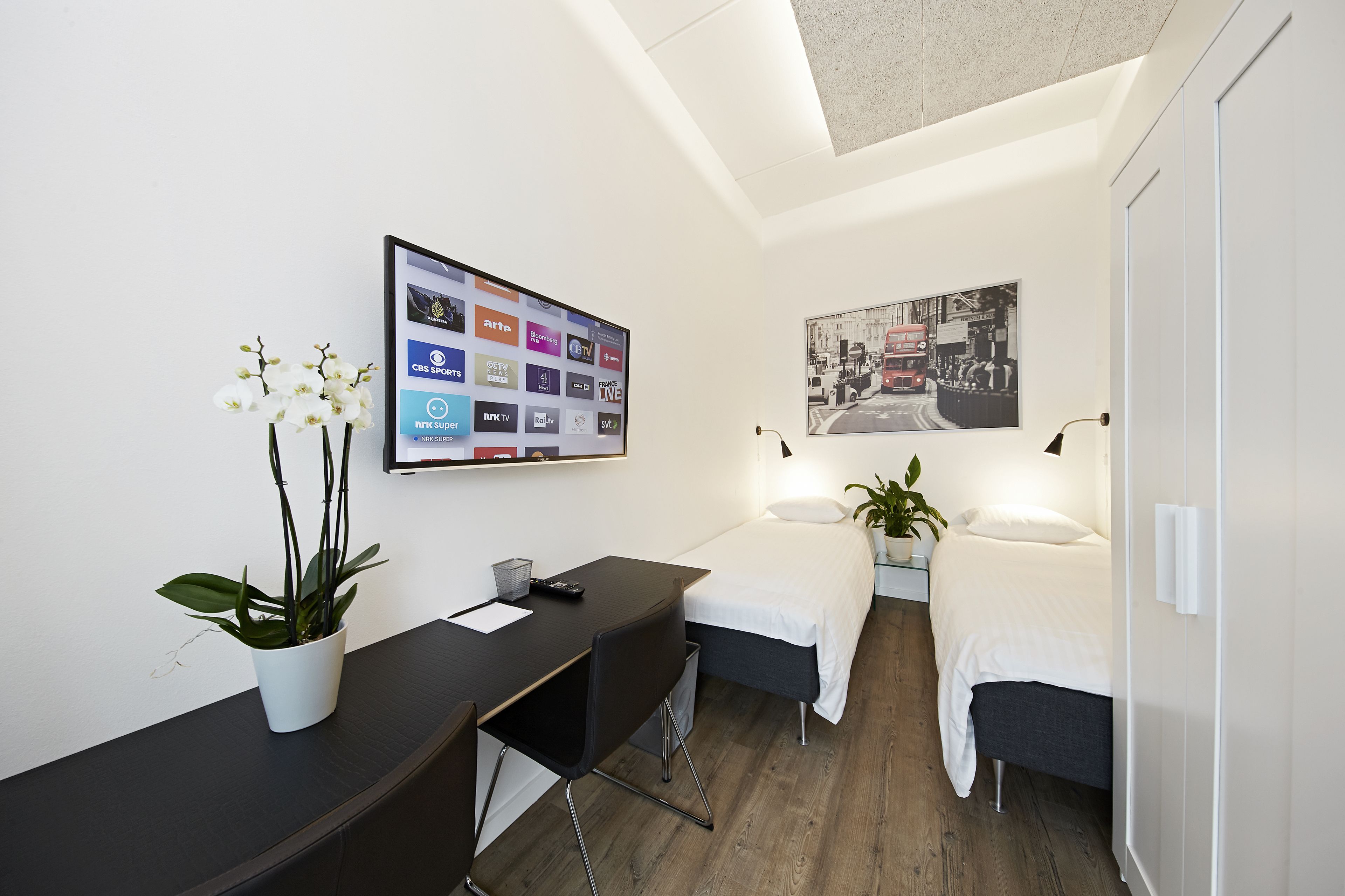 A modern hotel room with a desk, two beds, a flat-screen TV displaying channel logos, a framed picture on the wall, and a potted plant.
