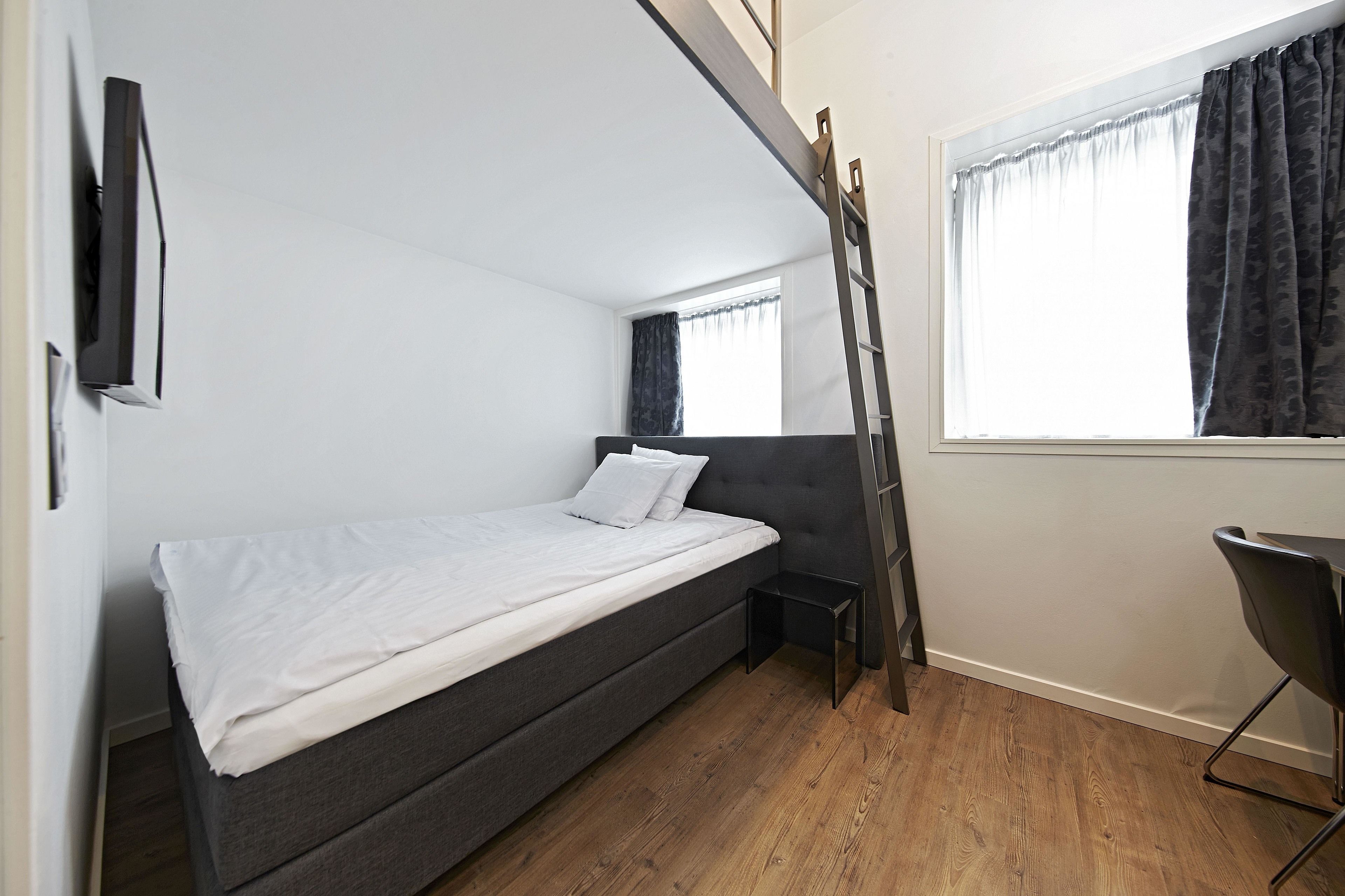 A compact room with a bed, a small table, a chair, a window with curtains, and a wooden floor.