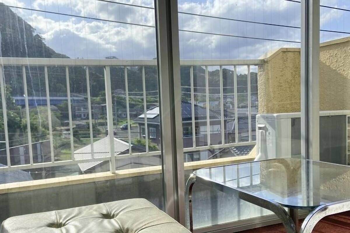 Een balkon met een glazen tafel en een gewatteerd zitje, met uitzicht op een woonwijk met huizen en een berg.