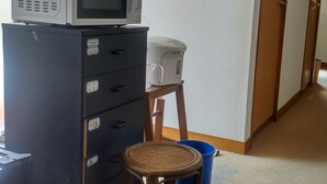 Une pièce avec un four à micro-ondes posé sur un meuble, un petit tabouret, une poubelle bleue et une porte à l’arrière-plan.