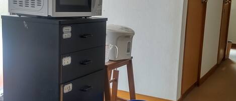 Une pièce avec un four à micro-ondes posé sur un meuble, un petit tabouret, une poubelle bleue et une porte à l’arrière-plan.