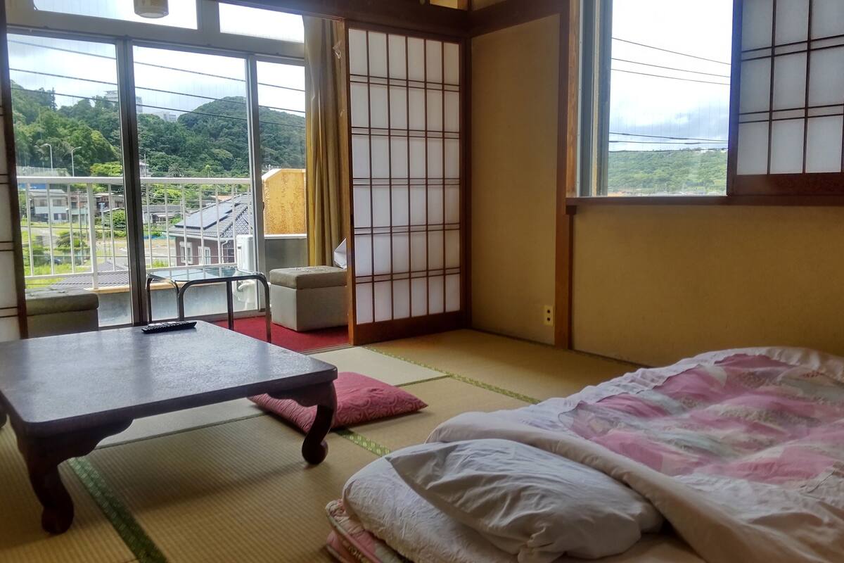 Een traditionele Japanse kamer met tatami vloerbedekking, een futonbed, een kleine tafel met een afstandsbediening, en een schuifdeur van shoji papier.