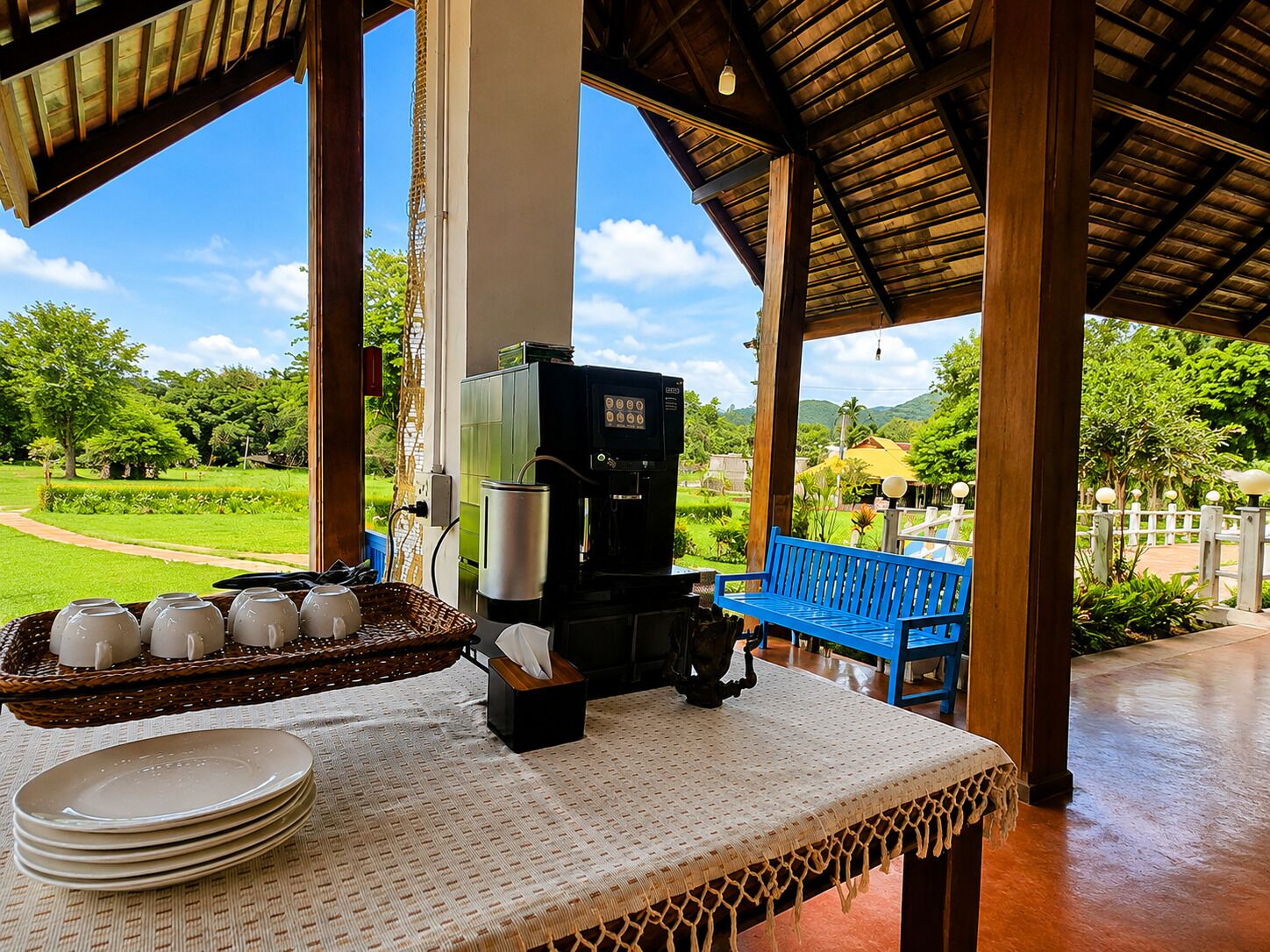 A covered outdoor seating area with a coffee machine, a table with plates and cups, and a view of a lush garden.