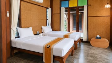 A hotel room with two beds, a wooden floor, an air conditioning unit, a bedside table with a tissue box, and a view through a window with curtains.