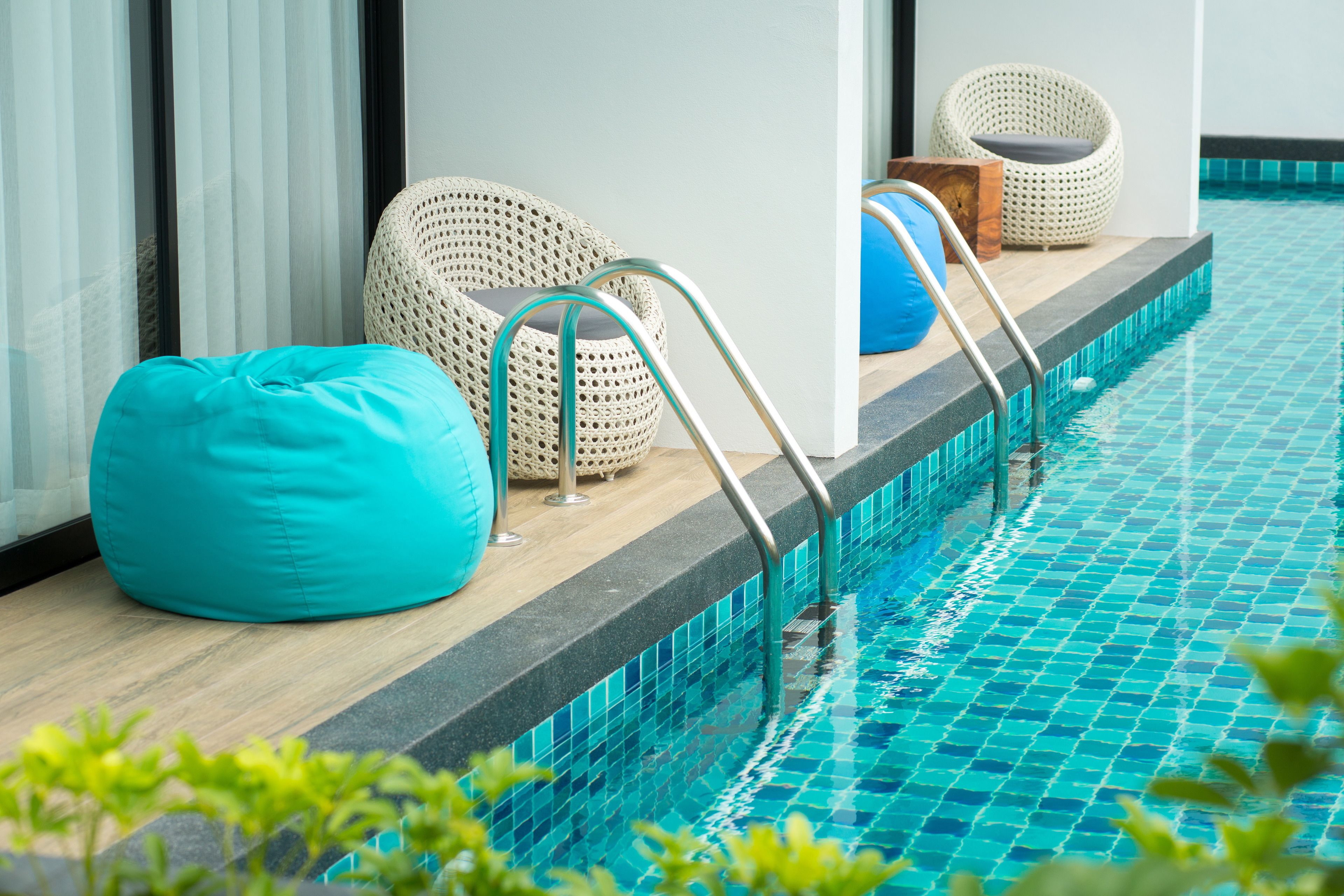 Un área junto a la piscina con un sillón tipo cojín azul, sillas de mimbre y un banco de madera.