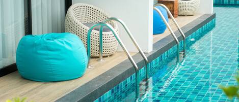 Un área junto a la piscina con un sillón tipo cojín azul, sillas de mimbre y un banco de madera.