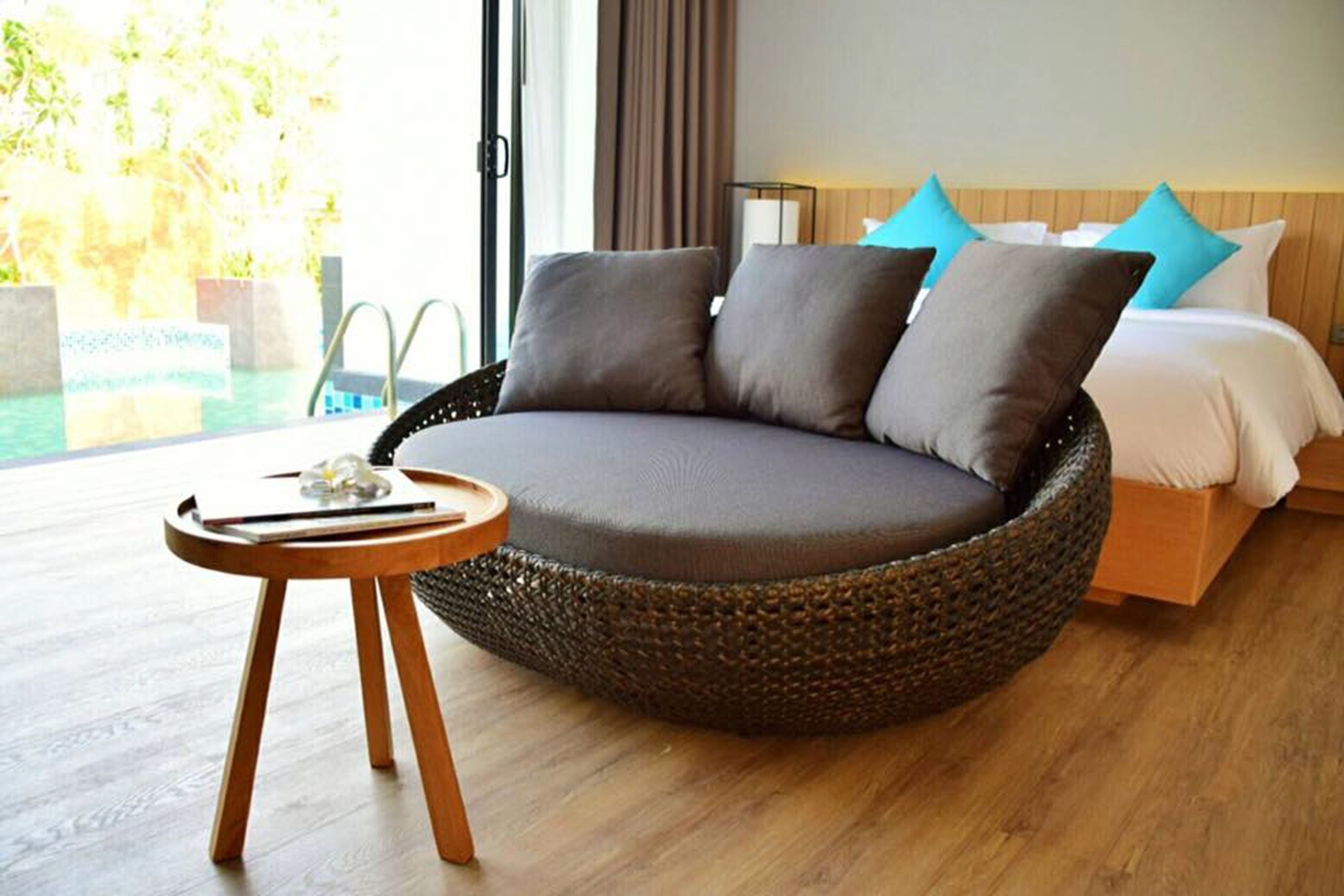 A modern room with a circular woven sofa, a small wooden side table, a bed with white and blue pillows, and a view of a swimming pool.