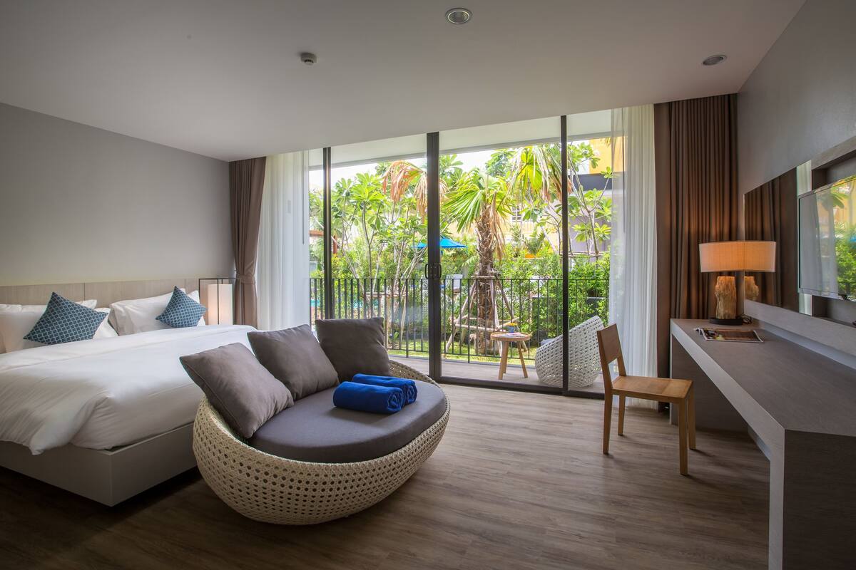 Quarto de hotel moderno com cama, área de estar com cadeira de fibra natural, escrivaninha e janela grande com vista para um jardim e palmeiras.