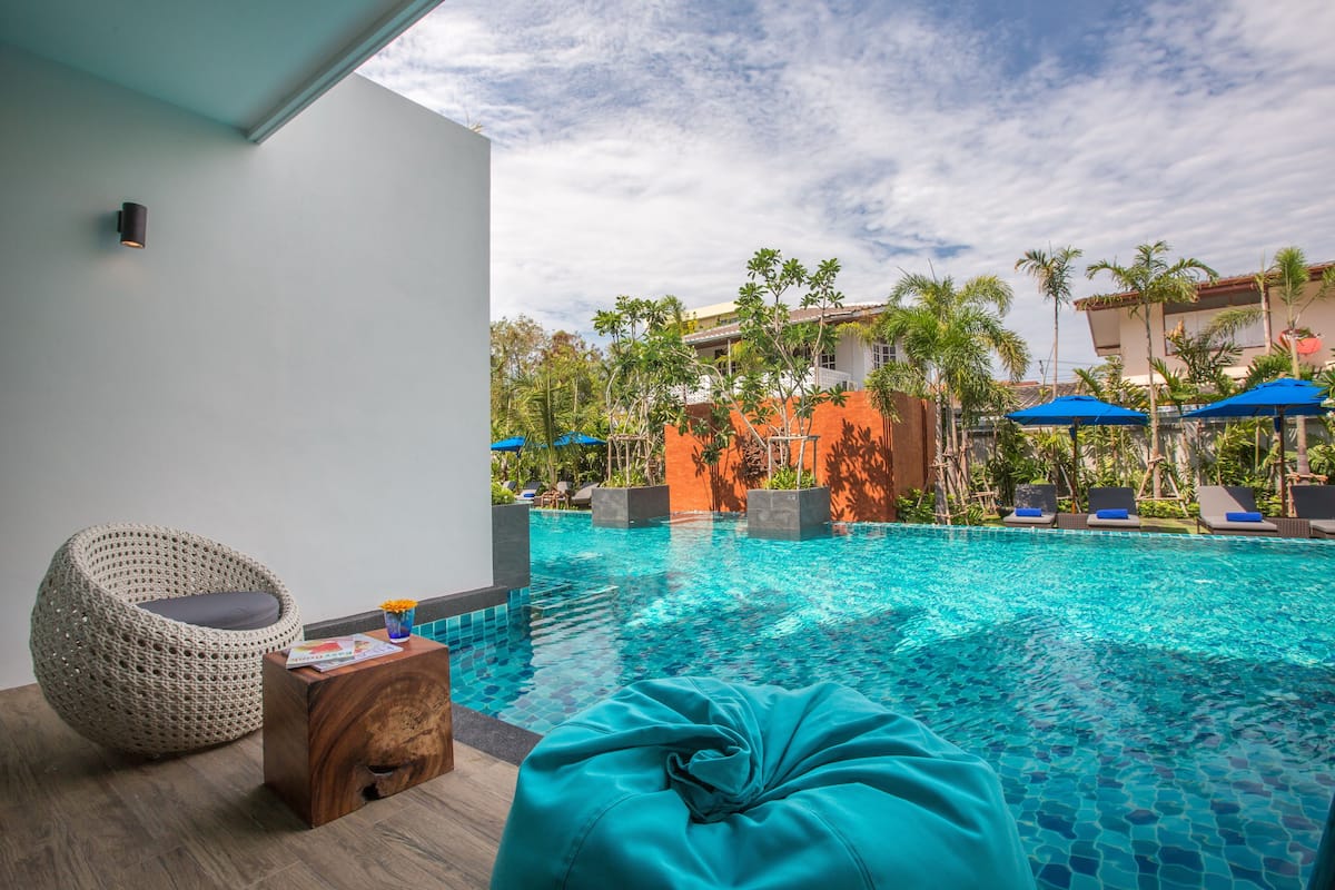 Uma área à beira da piscina com uma cadeira de vime, uma mesinha de madeira e um puff azul.