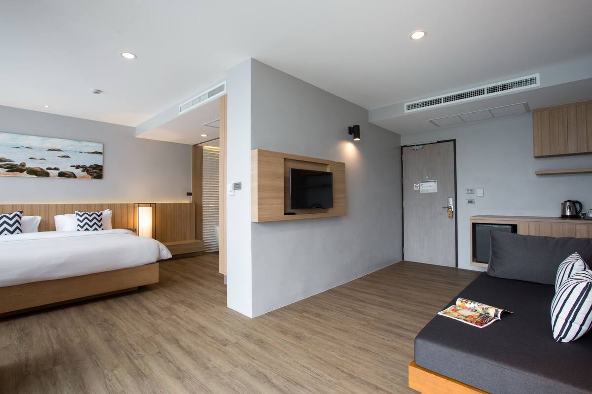 Quarto de hotel moderno com uma cama grande, um sofá menor, uma televisão de tela plana na parede, um armário de madeira e uma área de kitchenette.