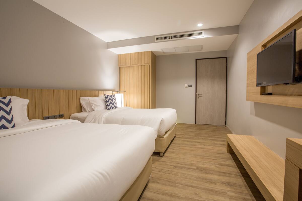 Quarto de hotel com duas camas, uma televisão, um banco de madeira e uma porta.