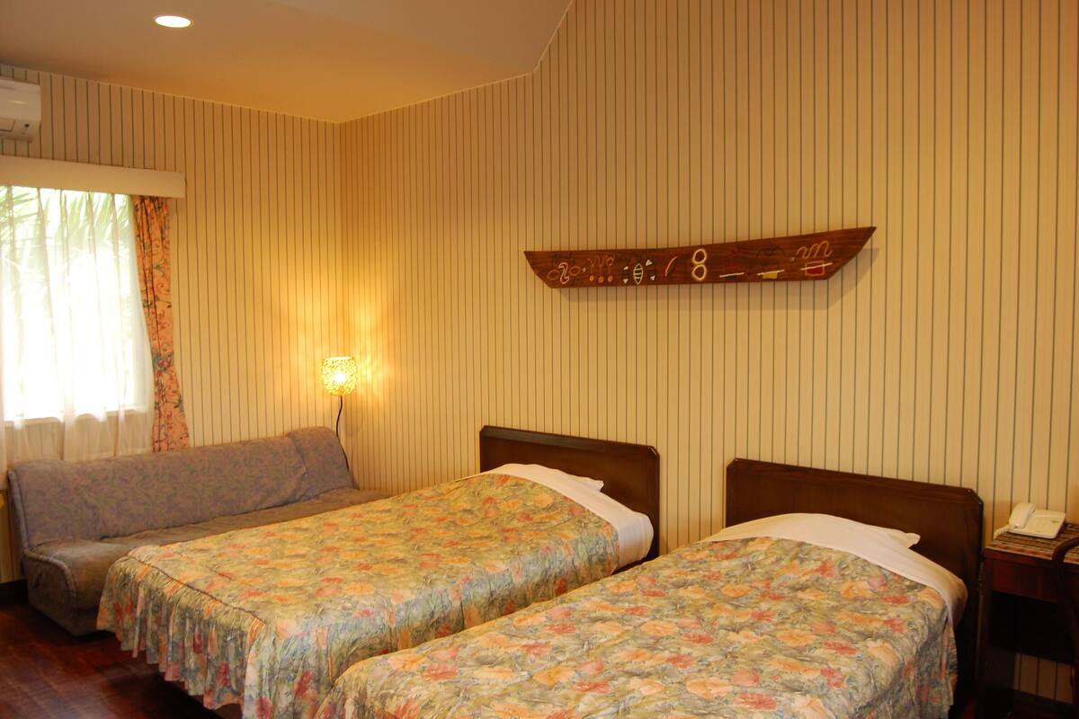 A hotel room with two single beds, a wooden headboard, a bedside table with a lamp, and a wall-mounted telephone.