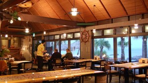 A restaurant with wooden tables and chairs, large windows with a view of the ocean, and a ship's wheel on the wall. - Amami Resort Basyayamamura (Amami)