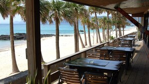 An outdoor seating area with tables and chairs overlooking a beach with palm trees and the ocean. - Amami Resort Basyayamamura (Amami)