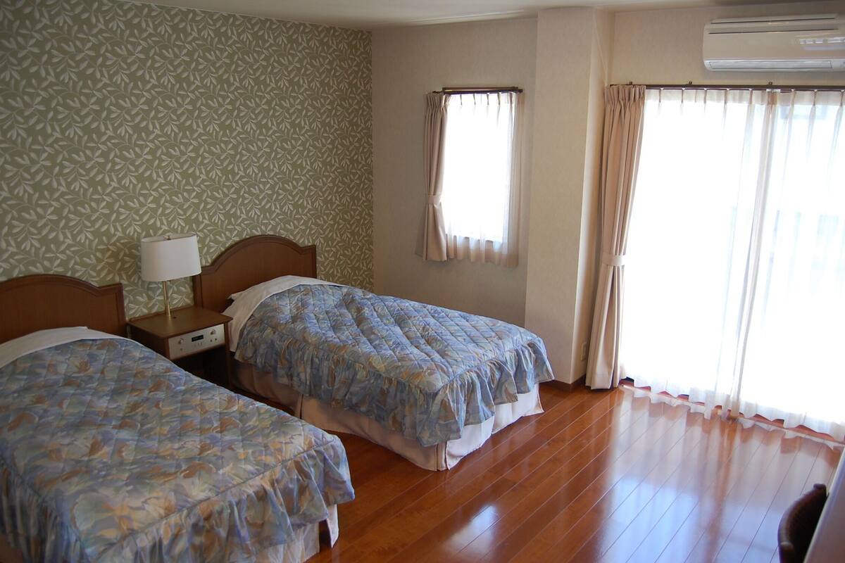 A hotel room with two beds, a wooden floor, patterned wallpaper, and a window with curtains.