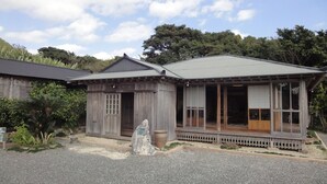 A traditional wooden house with a covered porch and a stone monument in front. - Amami Resort Basyayamamura (Amami)