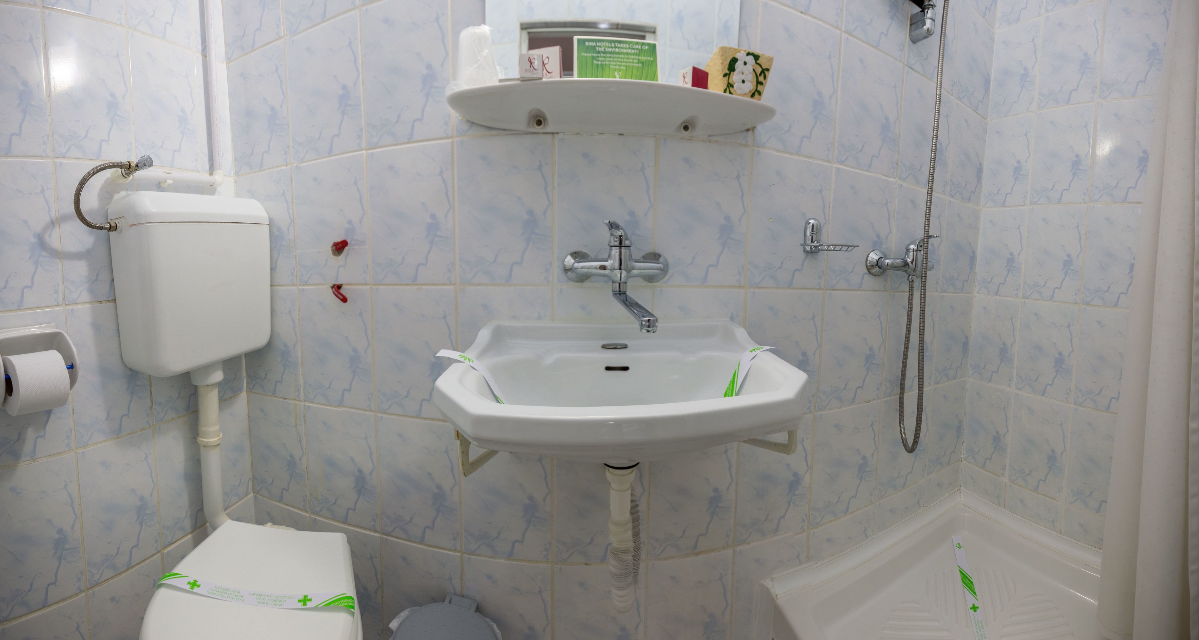 Une salle de bain comprenant des toilettes, un lavabo et une douche. Des étagères se trouvent au-dessus du lavabo, où sont rangés les produits de toilette.