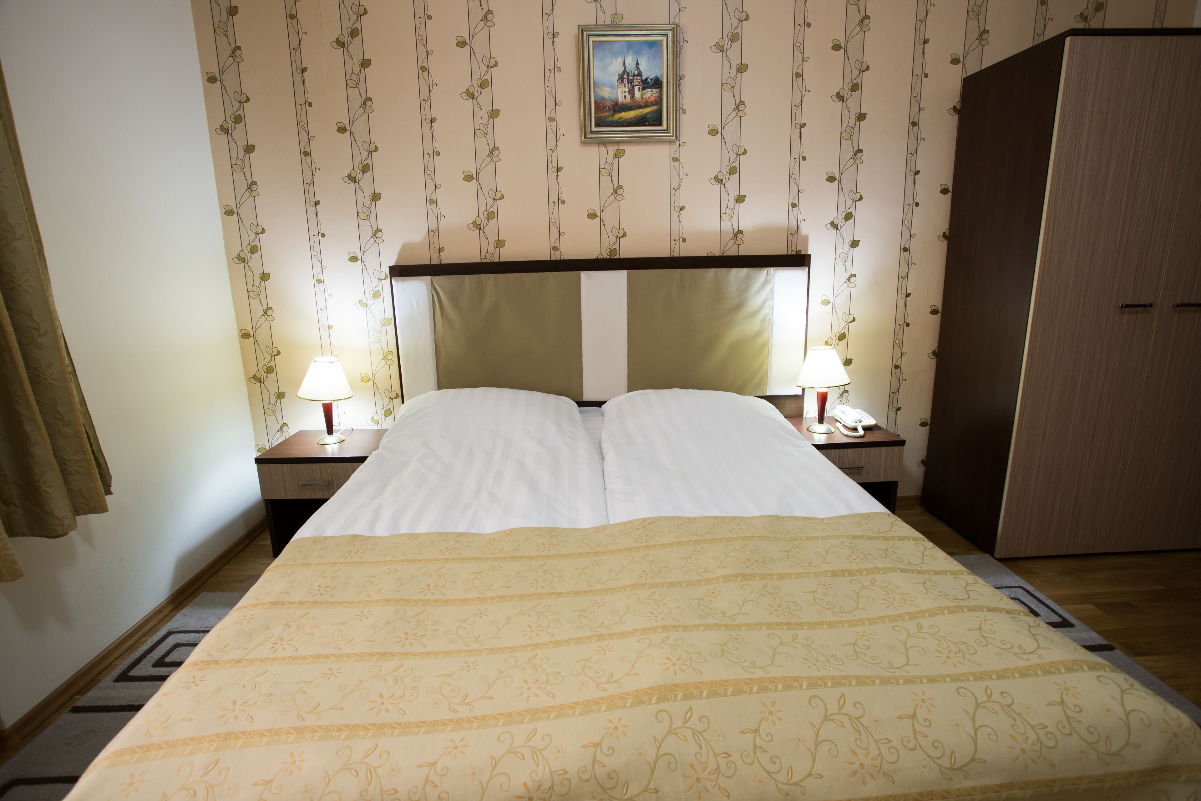 Una cama bien hecha con una colcha estampada, dos lámparas de noche, una mesita de noche, un armario y un cuadro en la pared.