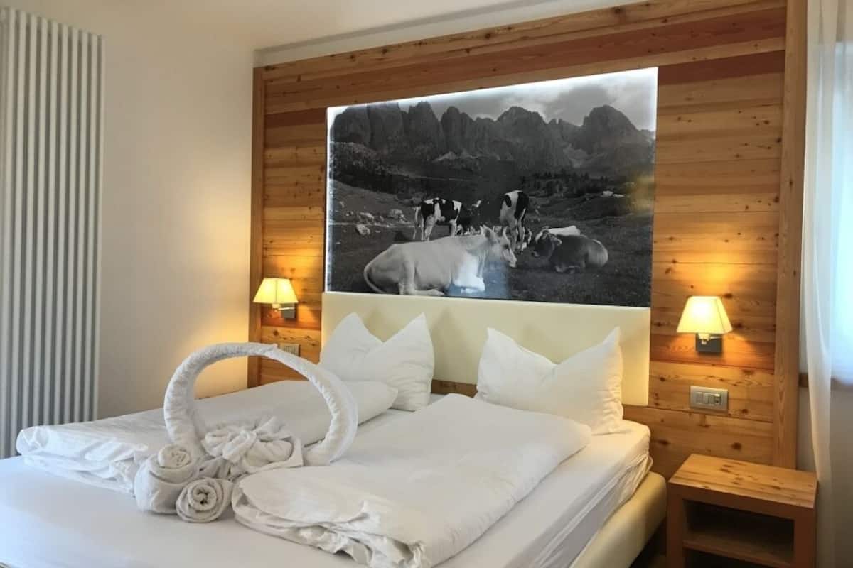 Ein Hotelzimmer mit einem hölzernen Kopfende, einem Bett mit weißer Bettwäsche, einer Handtuchgarnitur, zwei Nachttischlampen und einem großen Gemälde einer Berglandschaft.