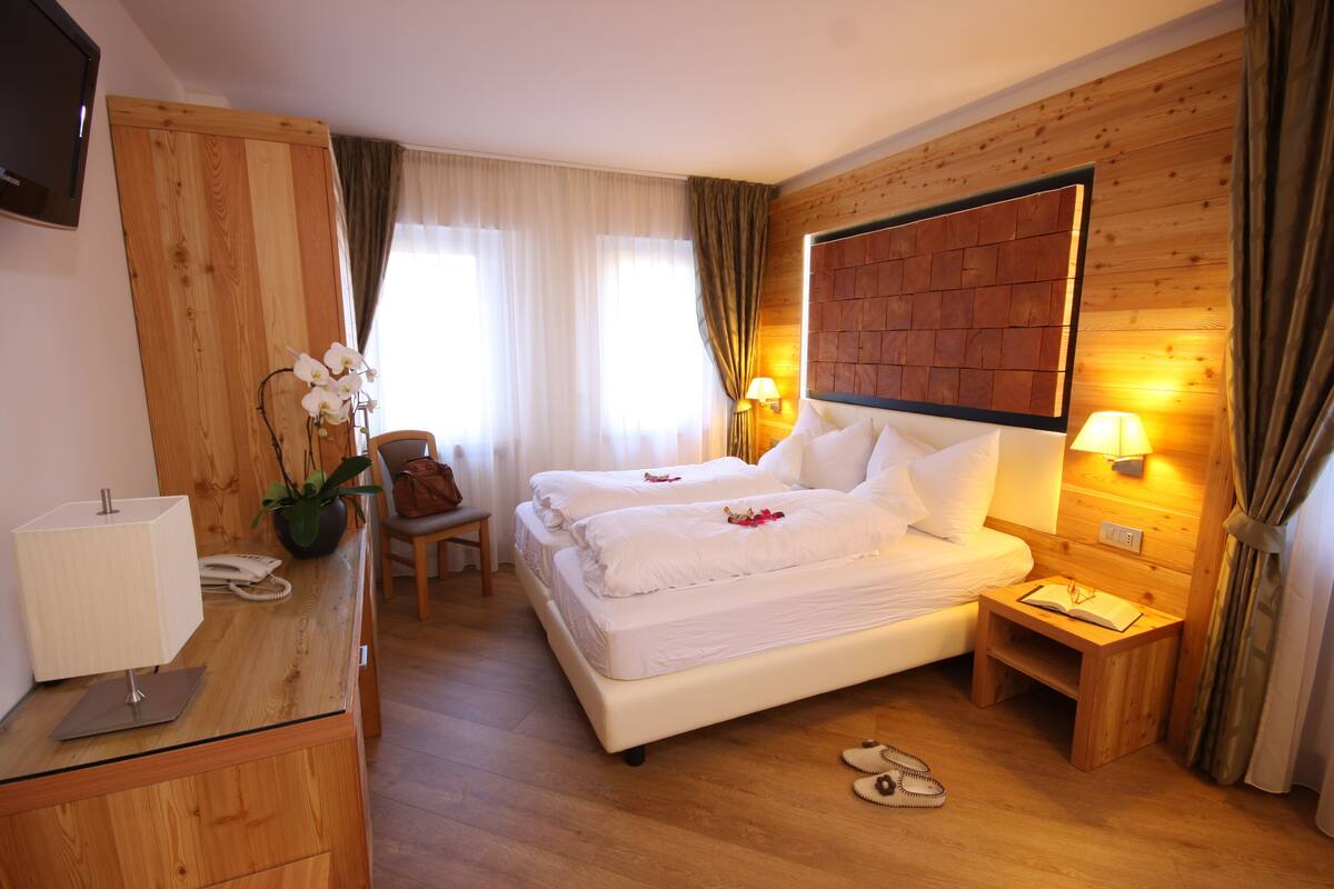 Ein Hotelzimmer mit einem hölzernen Kopfteil, zwei Betten, einem Schreibtisch mit Lampe, einem Sessel und einem kleinen Tisch mit einem Buch.