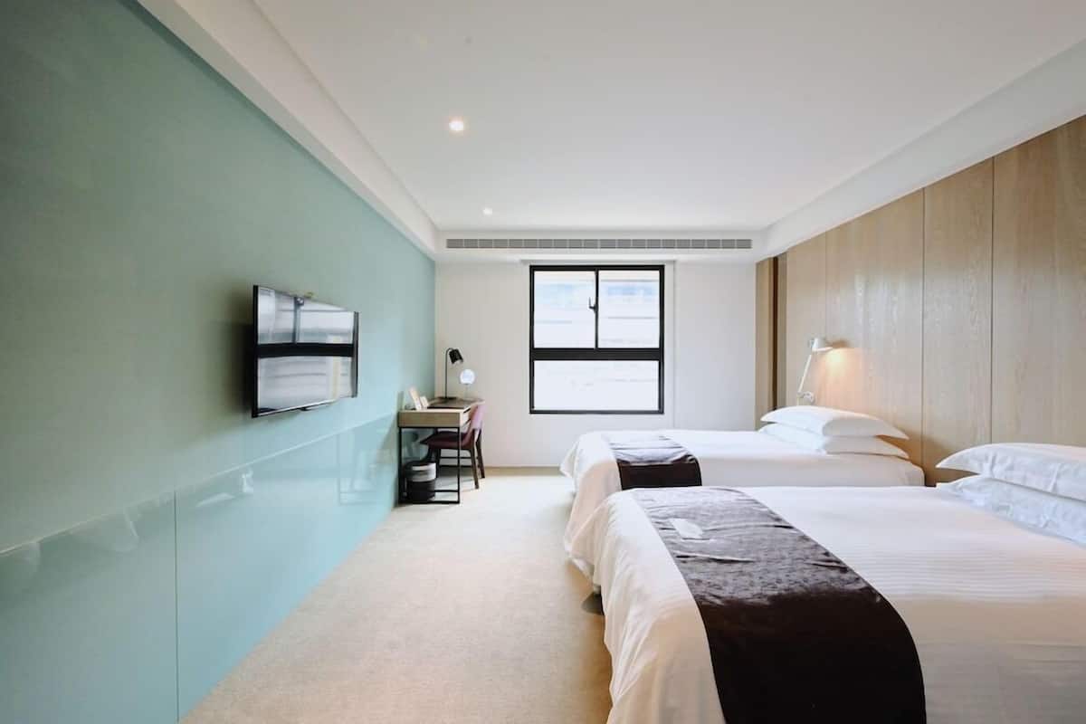 A hotel room with two beds, a desk, a chair, a TV, and a window.