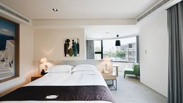 Habitación de hotel con una cama grande, dos mesitas de noche con lámparas, una silla verde, un escritorio y un ventanal con vista a la ciudad.