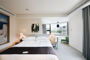 Habitación de hotel con una cama grande, dos mesitas de noche con lámparas, una silla verde, un escritorio y un ventanal con vista a la ciudad.