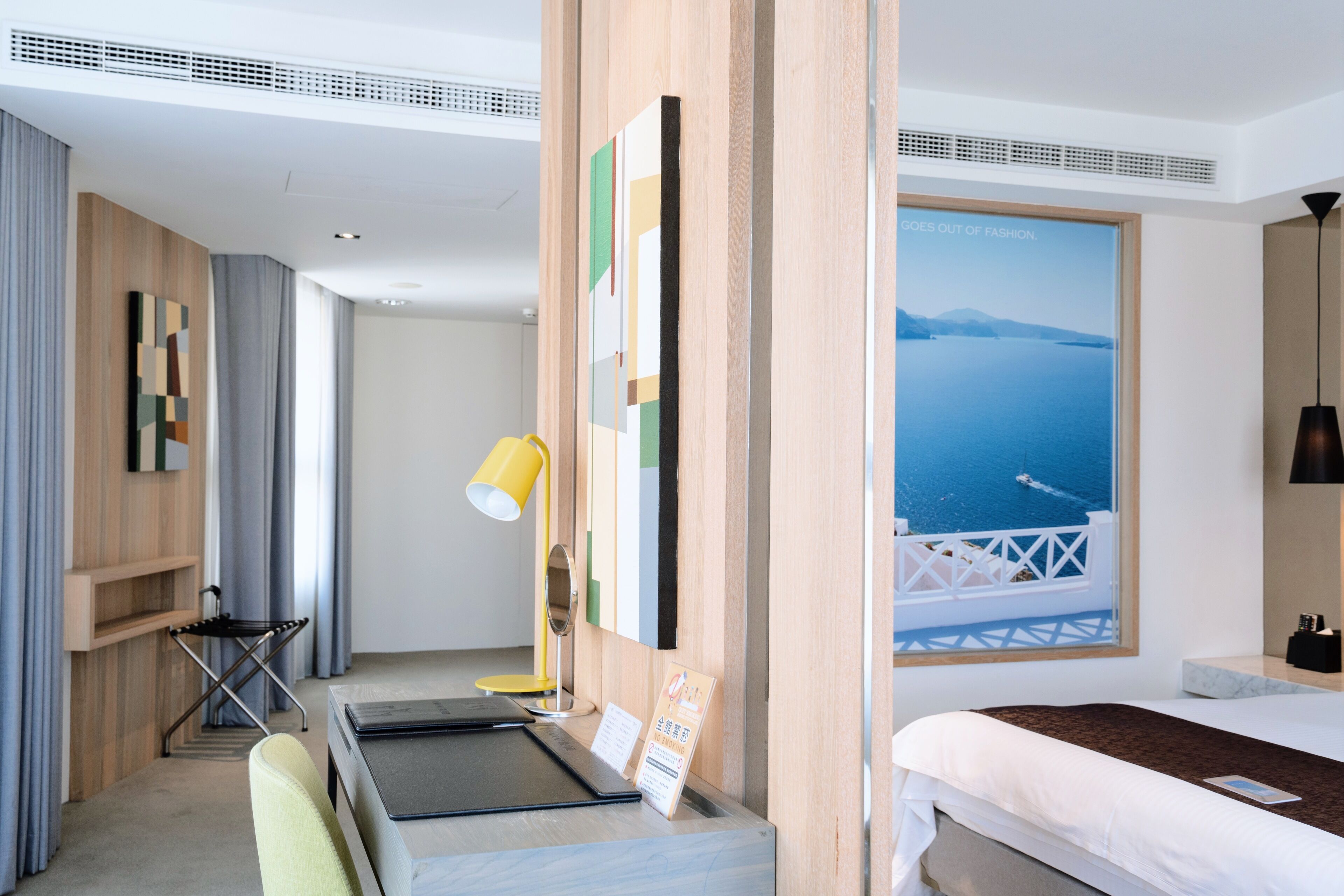 A hotel room with a bed, a desk, a chair, a lamp, and a view of the sea.