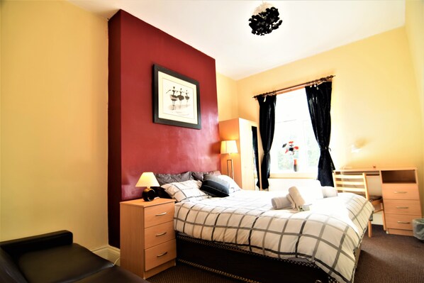 A bedroom with a bed, bedside tables, a desk, and a window with curtains. - Manu's Guesthouse (Birmingham)