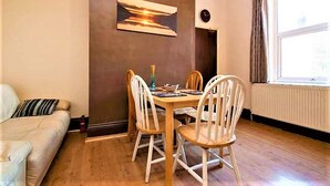 A dining area with a wooden table and four chairs, a painting on the wall, a sofa, and a window with curtains. - Manu's Guesthouse (Birmingham)