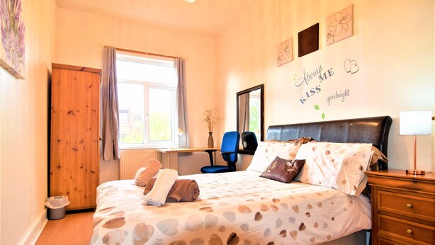 A bedroom with a bed, a desk, a chair, a dresser, and a window with curtains. - Manu's Guesthouse (Birmingham)