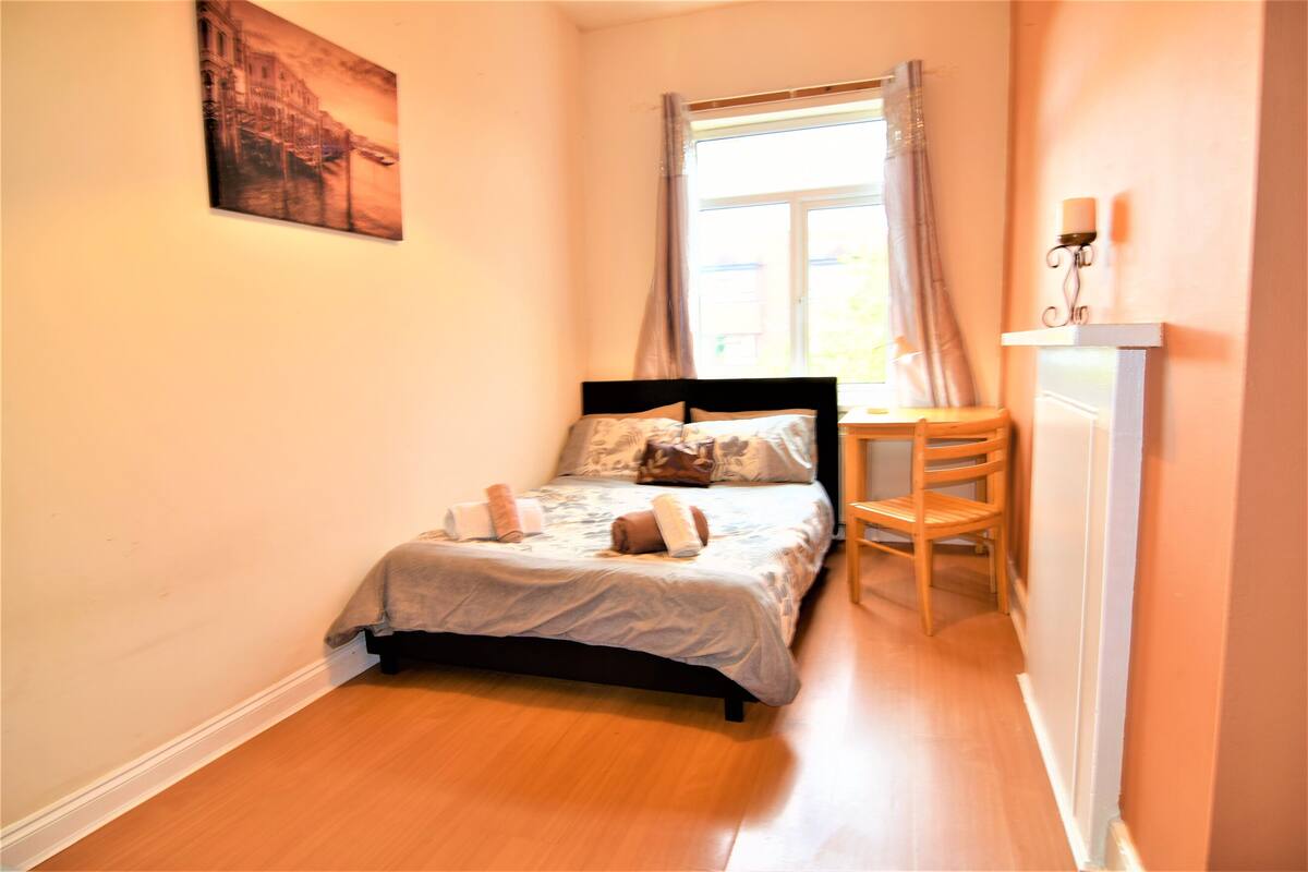 A narrow room with a bed, a chair, a window with curtains, and a wall-mounted light fixture.