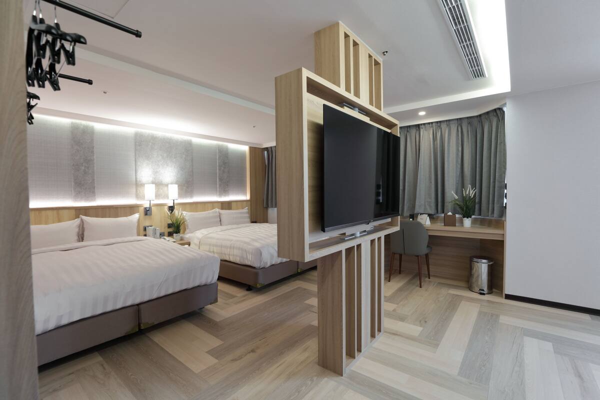 Une chambre d’hôtel moderne avec un grand lit, un téléviseur fixé sur une console en bois, un bureau avec une chaise et une petite poubelle.