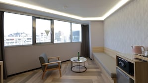 A modern hotel room with a large window, a small round table, a chair, a sofa, a kitchen area, and a city view. - Silken Hotel (Taipei)