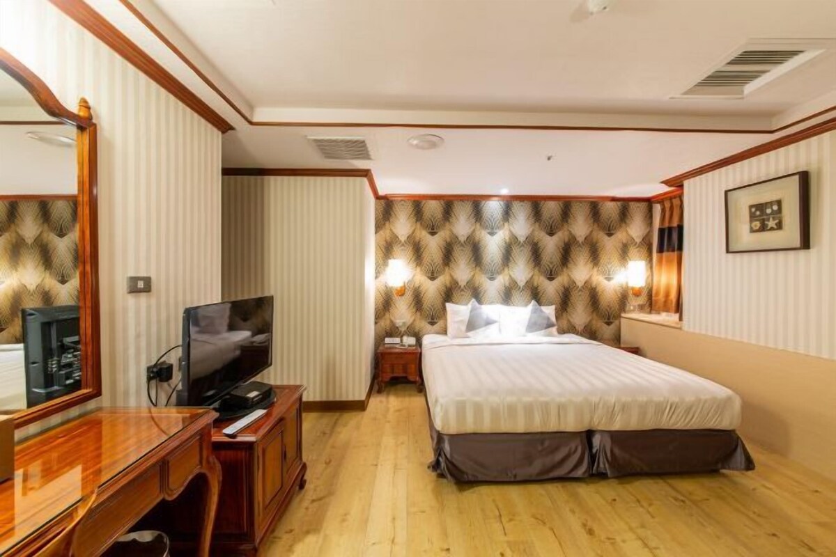 A hotel room with a wooden desk, a flat-screen TV, a large bed with a patterned headboard, and a wall-mounted lamp.