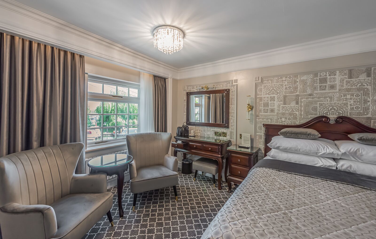 A hotel room with a bed, a desk, a chair, a window with curtains, and a patterned wall.