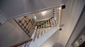 Uma escadaria com balaústres brancos e um corrimão de madeira, que leva a um quarto com um candelabro e um quadro na parede.